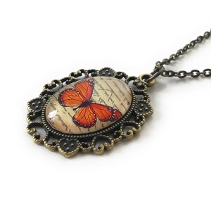 May include: A bronze necklace with an oval pendant featuring a vintage-style image of an orange butterfly on a cream background with cursive writing. The pendant is surrounded by a decorative filigree frame.