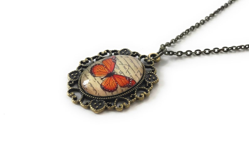 May include: A bronze necklace with an oval pendant featuring a vintage-style image of an orange butterfly on a cream background with cursive writing. The pendant is surrounded by a decorative filigree frame.