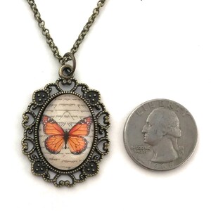 May include: A bronze necklace with an oval pendant featuring a vintage-style butterfly illustration on a cream background with handwritten text. The pendant is surrounded by a decorative filigree frame.