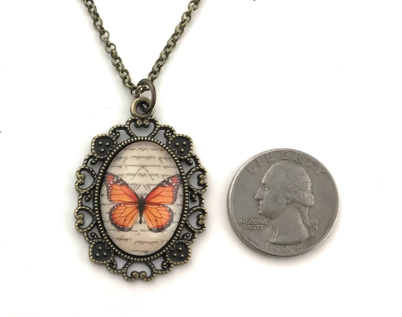 May include: A bronze necklace with an oval pendant featuring a vintage-style butterfly illustration on a cream background with handwritten text. The pendant is surrounded by a decorative filigree frame.
