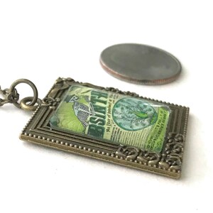 May include: A bronze pendant necklace featuring a vintage-style image of a skeleton holding a bottle of absinthe. The image is framed in a decorative bronze frame with a beaded edge. The pendant is about the size of a US quarter.