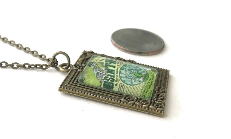 May include: A bronze pendant necklace featuring a vintage-style image of a skeleton holding a bottle of absinthe. The image is framed in a decorative bronze frame with a beaded edge. The pendant is about the size of a US quarter.