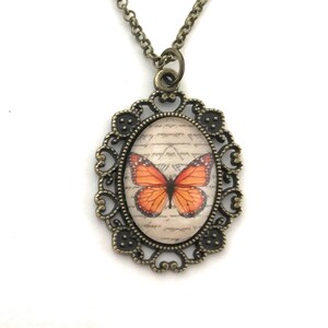 May include: A bronze necklace with an oval pendant featuring a vintage-style butterfly image on a background of handwritten script. The pendant has a filigree design.