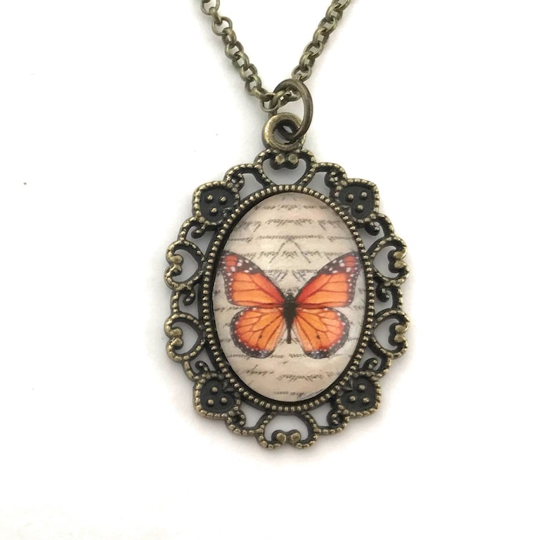 May include: A bronze necklace with an oval pendant featuring a vintage-style butterfly image on a background of handwritten script. The pendant has a filigree design.