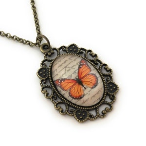 May include: A bronze necklace with an oval pendant featuring a vintage-style butterfly design on a background of handwritten script. The pendant is surrounded by an ornate filigree frame.