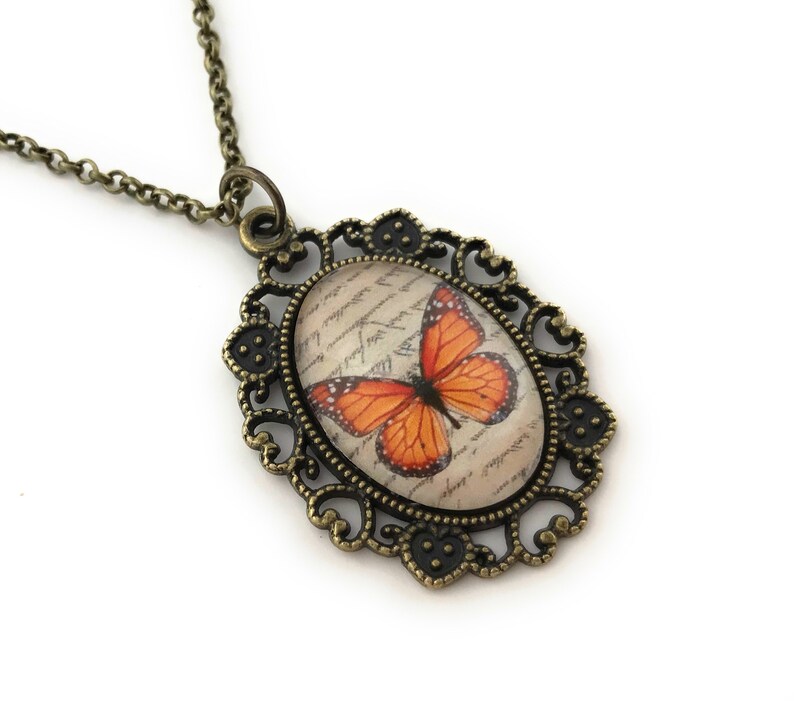 May include: A bronze necklace with an oval pendant featuring a vintage-style butterfly design on a background of handwritten script. The pendant is surrounded by an ornate filigree frame.