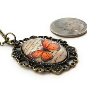 May include: A bronze necklace with an oval pendant featuring a vintage-style butterfly with orange wings on a cream background. The pendant is surrounded by a decorative filigree frame.