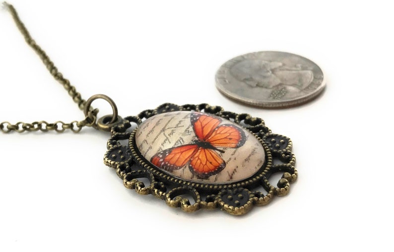 May include: A bronze necklace with an oval pendant featuring a vintage-style butterfly with orange wings on a cream background. The pendant is surrounded by a decorative filigree frame.
