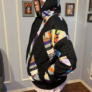 May include: A quilted black jacket with a hood, featuring colorful patchwork accents on the hood and sleeves. The jacket has a quilted pattern and a front zipper. The patchwork includes various colors and patterns.