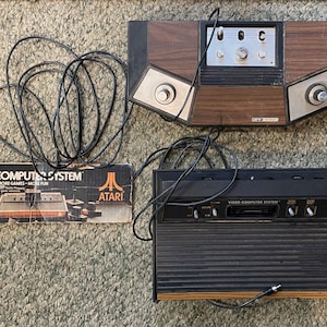 May include: Vintage Atari Video Computer System consoles and box. The consoles are black and brown with silver accents. The box has the words "Video Computer System" and "More Games - More Fun".