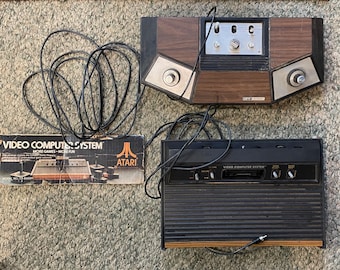 May include: Vintage Atari Video Computer System consoles and box. The consoles are black and brown with silver accents. The box has the words "Video Computer System" and "More Games - More Fun".