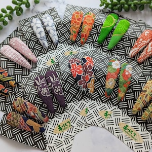 May include: A collection of colorful, patterned hair clips arranged on a black and white patterned surface. The clips feature various designs, including floral, geometric, and abstract patterns in shades of red, green, blue, and gold. Some clips have the text "手づくり HAND MADE".