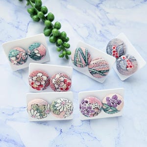 May include: Six pairs of fabric covered button earrings displayed on white cards. The earrings feature a variety of floral and geometric patterns in shades of pink, green, and purple.