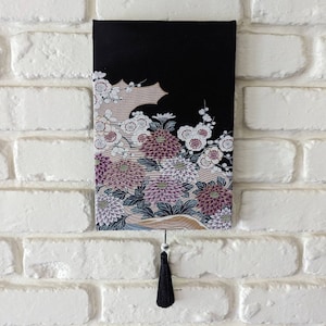 May include: A rectangular wall hanging with a black top and a floral design. The design features white, pink, and purple flowers with green leaves. A black tassel hangs from the bottom. The wall hanging is mounted on a white brick wall.