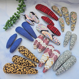 May include: An assortment of fabric-covered hair clips in various patterns and colors. The collection includes blue, red plaid, floral, and geometric designs. The clips are arranged on a light blue surface, showcasing a variety of styles.
