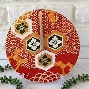 May include: A red, gold, and green clock with a Japanese-inspired design. The clock face is made of fabric with a geometric pattern. The clock hands are made of wood.