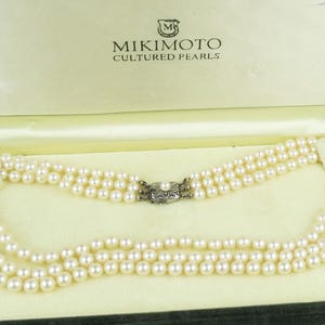 May include: A three-strand pearl necklace displayed in an open, dark green velvet box. The box is lined with a light green fabric and features the brand name "MIKIMOTO CULTURED PEARLS" printed on the inside.