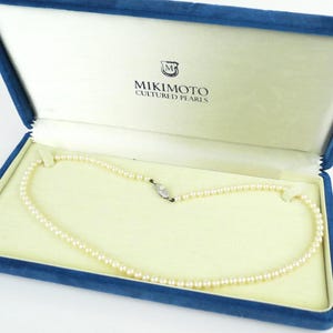 May include: A cultured pearl necklace by Mikimoto, presented in a blue velvet box. The necklace features small, round pearls and a silver clasp. The box has a cream interior and the Mikimoto logo is visible.