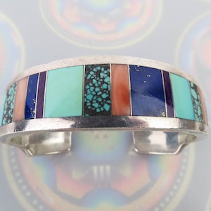May include: Silver cuff bracelet inlaid with rectangular sections of turquoise, coral, lapis lazuli, and other colorful stones. The bracelet has a polished finish and a classic Southwestern design.