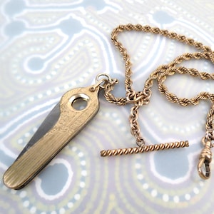 May include: A gold-toned pocket knife with an ornate design, paired with a matching gold chain. The knife is closed, revealing its decorative casing. The chain features a T-bar and a clasp. The items are set against a patterned background.