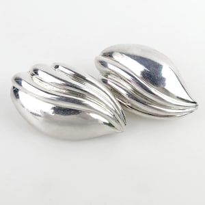 May include: Pair of silver-toned, leaf-shaped clip-on earrings. Each earring has a smooth, curved design with multiple ridges, creating a textured appearance. The earrings are displayed on a white background, highlighting their reflective surface.