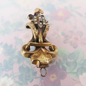 May include: Ornate antique gold-tone pendant with a floral design and small, clear gemstones. The pendant features intricate detailing, including scrolling patterns and a small loop at the bottom. The background is a soft pink with a floral pattern.