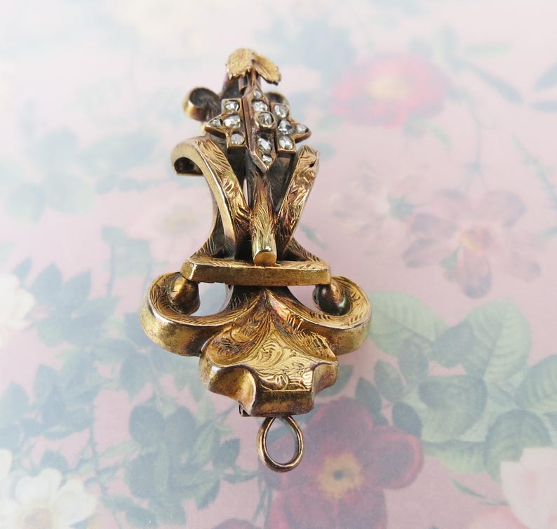 May include: Ornate antique gold-tone pendant with a floral design and small, clear gemstones. The pendant features intricate detailing, including scrolling patterns and a small loop at the bottom. The background is a soft pink with a floral pattern.