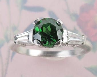 May include: A silver ring with a large, round, emerald-green gemstone at its centre. Two smaller, rectangular, clear gemstones are set either side of the main stone. The ring has a classic, elegant design.