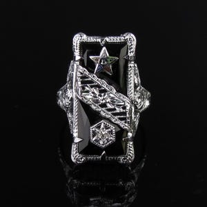 May include: A silver-toned ring with a rectangular black onyx center stone. The ring features a star and geometric designs, with intricate detailing on the sides. The ring has a vintage or antique aesthetic.