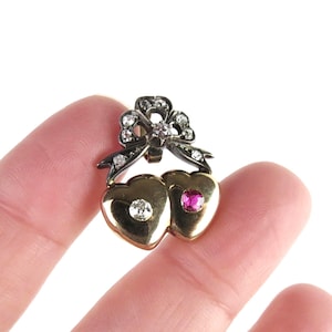 May include: A vintage brooch featuring two gold hearts, each adorned with a gemstone. One heart has a clear stone, the other a pink stone. A bow with clear stones sits above the hearts. The brooch is held by a finger.