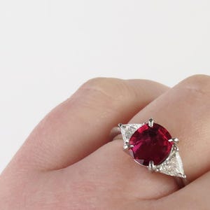 May include: A silver ring featuring a large, oval-cut, deep red gemstone, flanked by two triangular, clear gemstones. The ring is set against a neutral background, highlighting the vibrant color of the central stone.