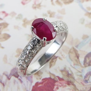 May include: A silver ring featuring a large, oval-cut ruby gemstone. The ruby is a deep red color and is set in a four-prong setting. The ring's band is encrusted with small, clear gemstones. The ring is displayed on a floral background.