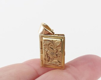 May include: A small, gold-colored locket pendant with a floral design. The rectangular pendant opens to reveal a small space. The front of the locket features an engraved flower and leaf pattern. The pendant is held by a finger.