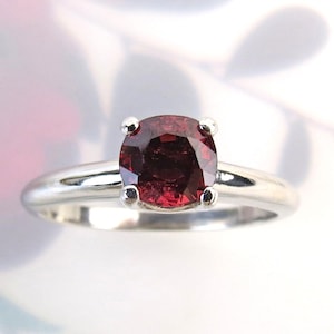 May include: A silver ring featuring a round, deep red gemstone. The gemstone is the focal point, set with four prongs on a polished silver band. The ring is displayed against a soft, blurred background.