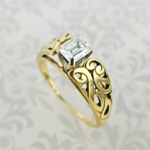 May include: A gold ring featuring an emerald-cut clear gemstone set in the center. The band is crafted with an openwork filigree design that extends from the setting down the sides of the ring, showcasing intricate swirling metal patterns.