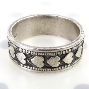 May include: A silver-toned metal band ring featuring a repeating pattern of embossed hearts around the center. The design is bordered by two thin, textured rope-like bands, with a dark, oxidized finish highlighting the heart shapes and details.