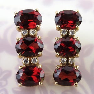 May include: A pair of gold earrings featuring three oval-cut, deep red gemstones. Small, clear accent stones are set between the red gems. The earrings have a classic, elegant design.