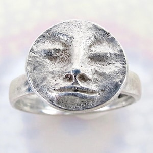 May include: A silver ring featuring a detailed, textured face design. The face has closed eyes, a small nose, and a subtle smile. The ring band is smooth and simple, complementing the detailed face.