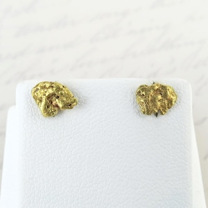 May include: A pair of gold-colored stud earrings. Each earring has a textured, irregular shape, resembling a small gold nugget. The earrings are displayed on a white surface, with a soft, blurred background.