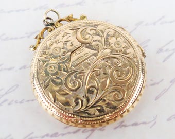 May include: A round gold locket featuring intricate floral and scroll engravings across the surface. The locket includes a small decorative bail at the top and a central cartouche displaying engraved hallmarks, including the text 9 375.