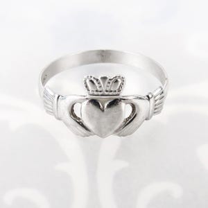 May include: A silver-toned Claddagh ring featuring a central heart held by two hands, topped with a crown. The metal band is smooth and circular, showcasing a polished finish that reflects light across the heart, hands, and crown design elements.