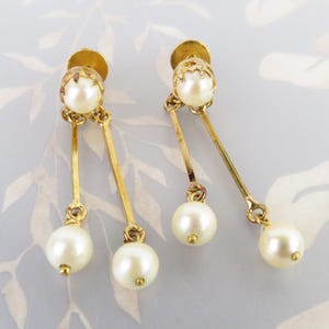 May include: A pair of vintage gold-tone earrings featuring faux pearls. Each earring has a pearl stud, a gold bar, and a dangling pearl. The earrings are set against a soft, patterned background.