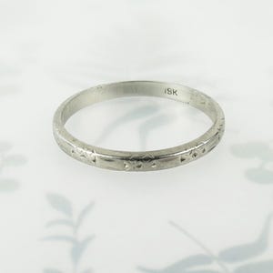 May include: A thin silver-toned metal band ring featuring a repeating engraved geometric pattern along the outer surface. The interior of the band is clearly stamped with the text 18K, indicating the metal purity of this circular jewelry piece.
