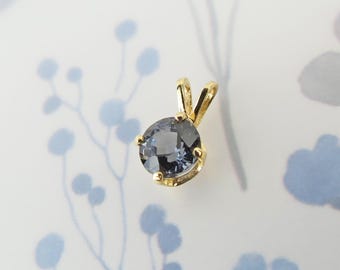 May include: A gold pendant with a round, gray-blue gemstone. The pendant has a simple, elegant design with a small loop for a chain. The background is a light blue and white floral pattern.