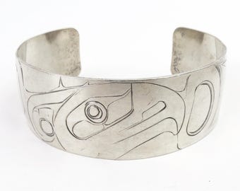 May include: A silver cuff bracelet with an engraved Native American design. The bracelet features a detailed carving of a bird or animal, with intricate lines and shapes. The open-ended design allows for easy wear.
