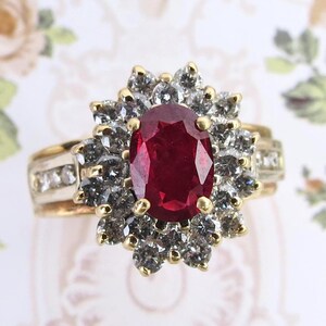 May include: A gold ring featuring a large, oval-cut deep red ruby at the center, surrounded by a halo of numerous small, round, clear white diamonds. The band is also set with a row of smaller diamonds along each side of the central cluster.