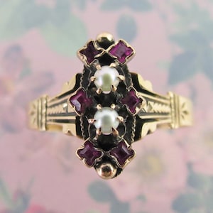 May include: A vintage gold ring featuring a central design with two pearls and four square-cut, deep red gemstones. The ring has an ornate, filigree-style setting with a black background, and is set on a gold band.