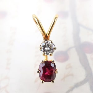May include: A gold pendant featuring a round, clear, faceted gemstone positioned above an oval, deep red, faceted gemstone. The pendant includes a V-shaped gold bail at the top for attachment, with both stones secured in gold prong settings.