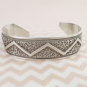 May include: A silver cuff bracelet with a textured band and a zig-zag design. The bracelet has an open design and is made of a shiny metal. The band features a detailed pattern and a geometric design.