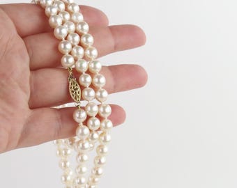 May include: A three-strand pearl necklace with a gold clasp. The pearls are a creamy white colour and appear to be round. The clasp is ornate and has a filigree design. The necklace is held in a hand against a white background.